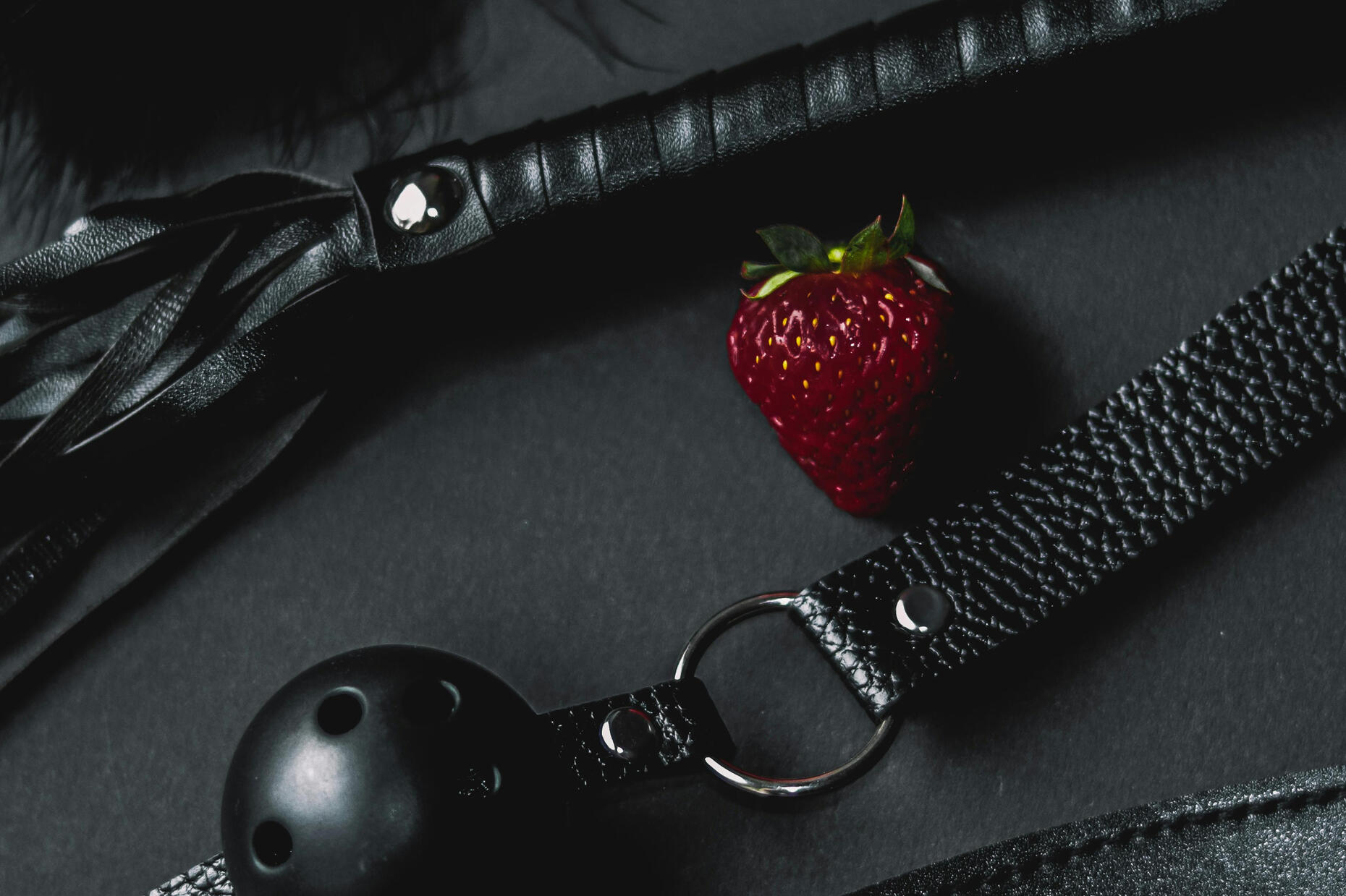 A flatlay of black BDSM gear set against a dark background. The composition includes a flogger with multiple leather strands, a textured leather strap with silver hardware, a perforated black ball gag, and a smooth blindfold or restraint. In the center of