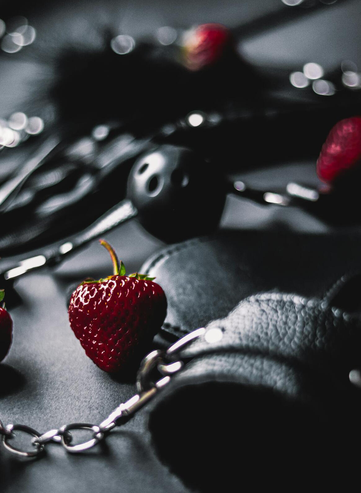 A moody close-up of BDSM gear and strawberries arranged on a black surface. In the foreground, two bright strawberries sit beside a pair of padded leather cuffs connected by a silver chain. In the blurred background, more strawberries and gear — including
